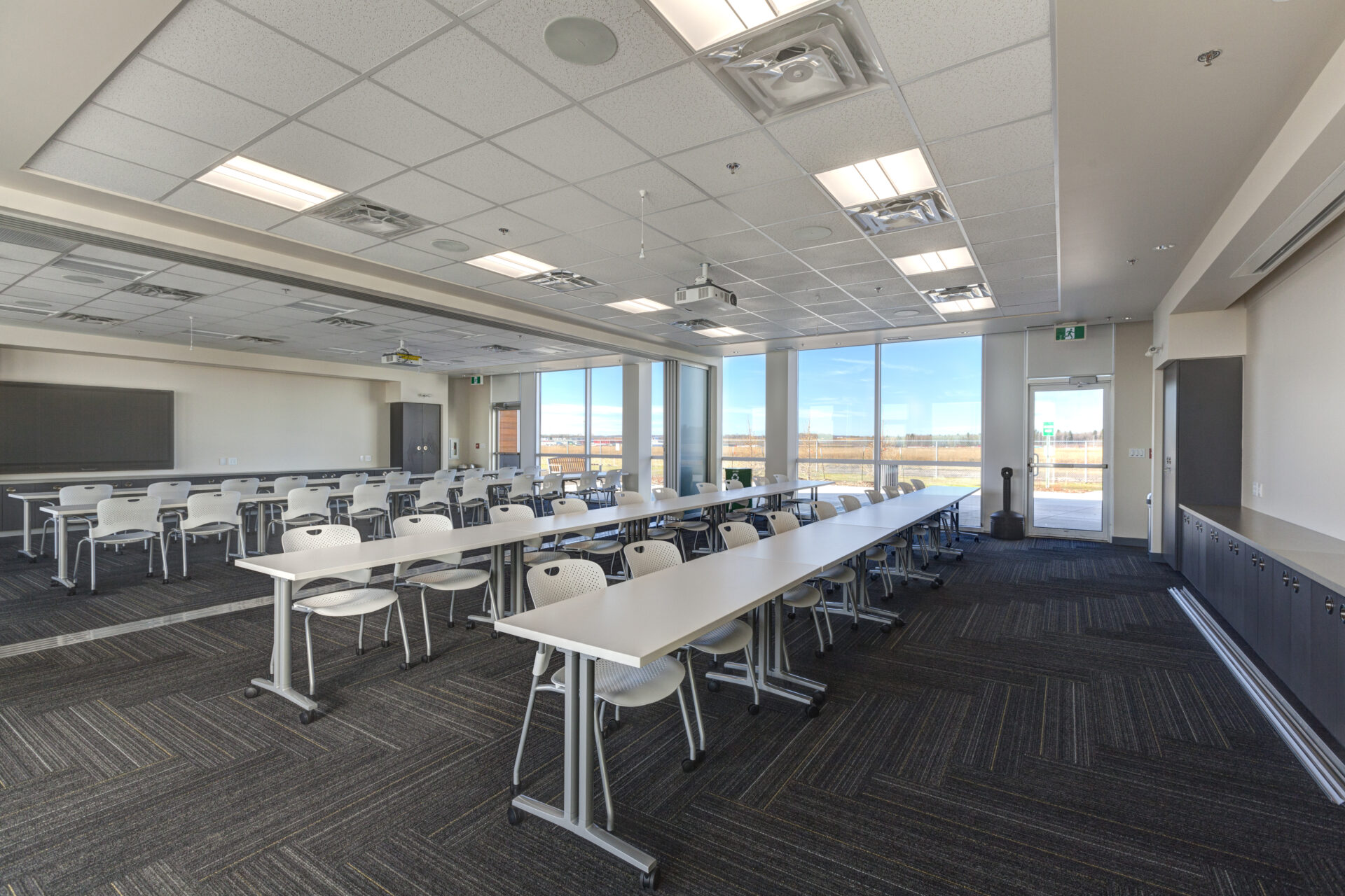 Large meeting room fitted with long white tables and black chairs. The room is brightly lit because of the floor to ceiling windows overlooking the landscape.