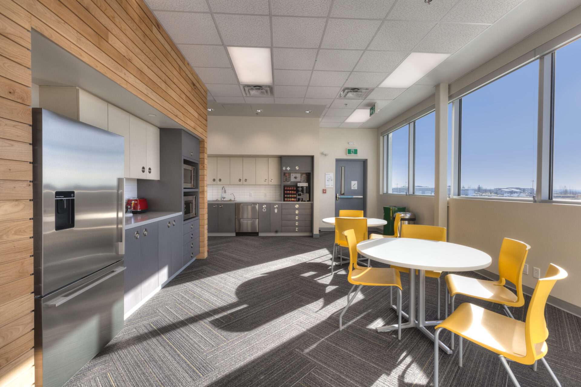Brightly commercial office kitchen space. Grey and off white cabinets on the left and back walls. Stainless steel appliances. Dark grey herringbone patterned carpet. Two smaller round tables with yellow accent chairs.