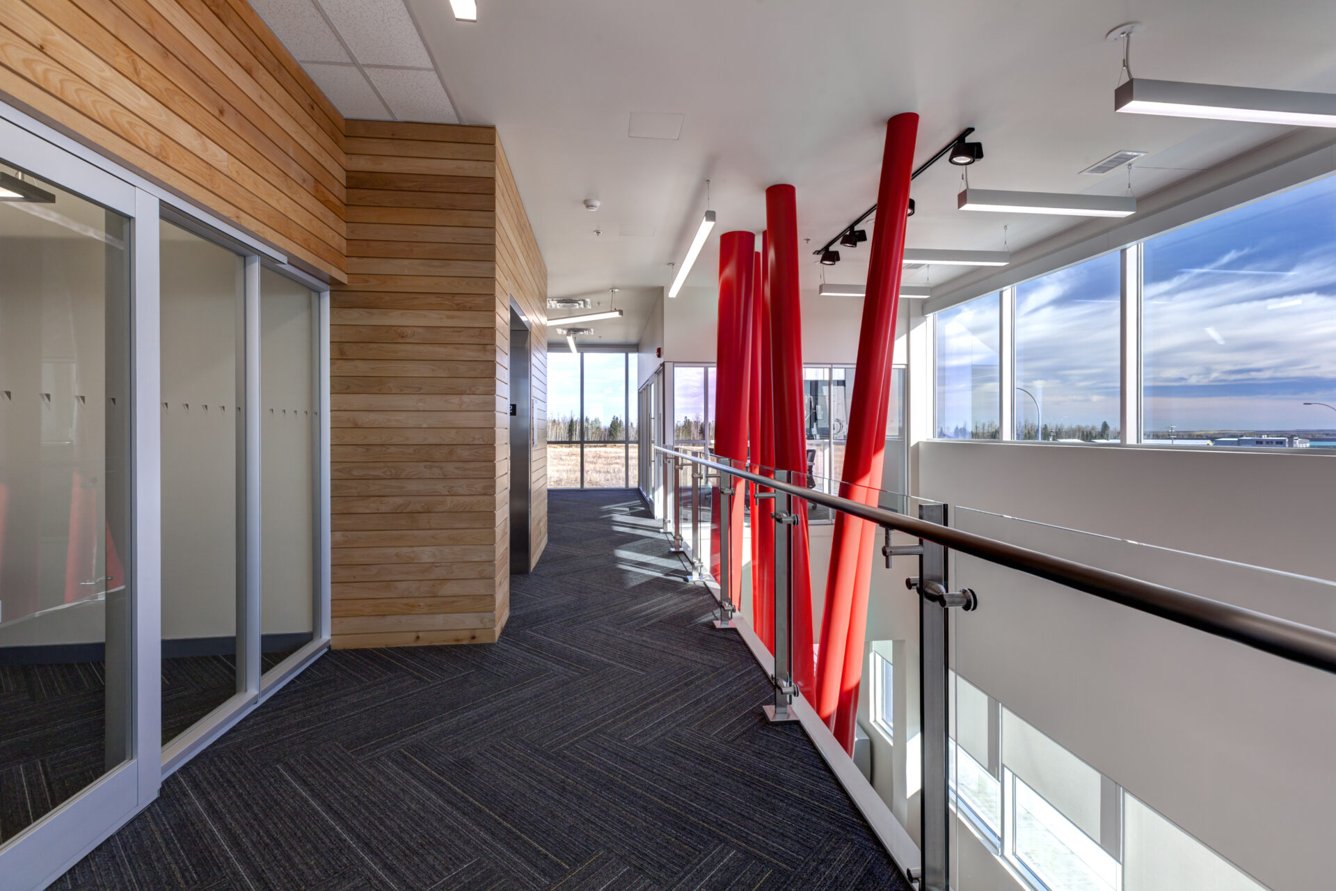 Second floor hallway looking into the atrium that features floor to second ceiling, red structural beams. It is brightly lit from the windows on main and second level. Dark grey herringbone patterned carpet. Horizontal wood slats, natural colour, on the left walls. Bright white walls and ceilings.