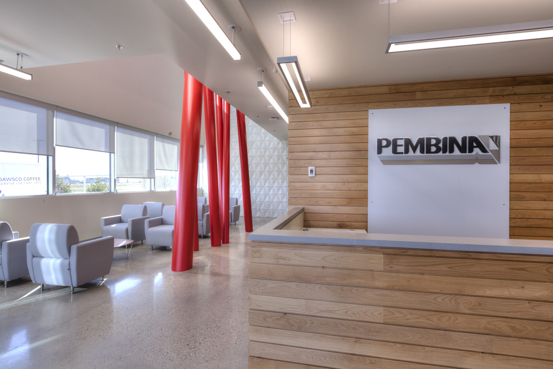 Bright reception area featuring grey chairs in the background and a bright window. In the middle ground there are red structural beams extended from ground to ceiling, a common visual piece throughout the building. In the foreground there is the reception desk fitted with horizontal wooden slats with the Pembina logo featured on the back wall.